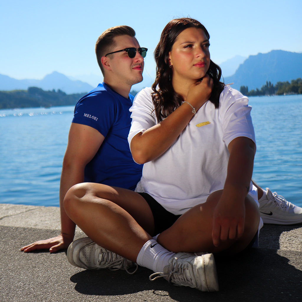 Ein Mann und eine Frau sitzen am Ufer. Der Mann trägt ein Navy Blaues T-Shirt mit weissem Farbfleck die Dame trägt ein Weisses Oversize T-Shirt mit goldenem Farbfleck