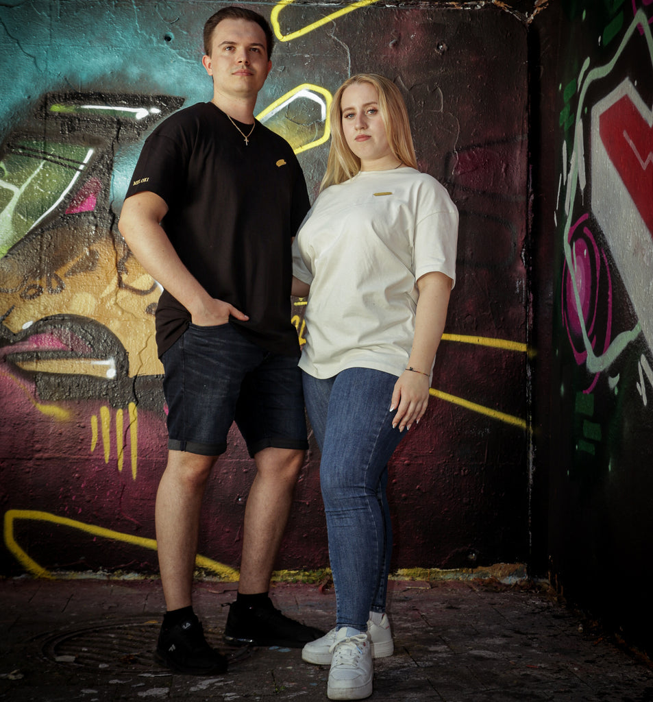 Ein Mann und eine Frau stehen vor einer Wand voller Graffitis. Sie tragen die Meloki Oversize T-Shirts mit goldenem Farbfleck